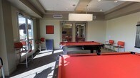 This single-floor recreational space is thoughtfully designed to serve as a social hub for games and gatherings. The room's most prominent feature is a full-size pool table dressed in bold red felt, anchored beneath a rectangular pendant light fixture that provides focused overhead illumination. The tray ceiling adds architectural depth to the space, while recessed lighting supplements the natural light that streams in through large windows along one wall, casting bright patches across the carpeted floor.

The room is accented with red and navy furnishings, including bar-height chairs and a compact mini-fridge tucked into a corner, offering convenience for extended leisure sessions. A framed red artwork on the wall ties the color scheme together cohesively. Glass-paneled double doors connect this game area to an adjacent room, visible through the panes, which appears to serve as a lounge or meeting space outfitted with additional red seating and tables. The open sightlines between the two areas create a sense of continuity, making the overall layout well-suited for hosting social events and casual recreation.