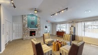This single-floor clubhouse offers a well-appointed community gathering space designed to accommodate a variety of social functions. The main lounge area serves as the centerpiece, featuring a dramatic floor-to-ceiling stone fireplace set against a teal accent wall, vaulted ceilings with ceiling fan, and track lighting throughout. Comfortable striped armchairs arranged around natural wood stump side tables create an inviting seating arrangement. French doors and large windows allow ample natural light to fill the space.

Adjacent to the lounge, open shelving units display decorative accents, while a dedicated dining area includes bar-height seating along a counter-height table paired with a teal bistro table and chairs. A separate gathering room provides additional flexible seating with carpet flooring and multiple windows.

The fully equipped kitchen features dark cabinetry contrasted with light granite-style countertops, stainless steel appliances including a range and microwave, and a large island with bar stool seating. A sideboard and beverage station near the lounge offer convenient refreshment access. The overall ambiance balances casual comfort with functional design, making it suitable for both quiet gatherings and larger community events.