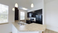 Located at 203 Magnolia Blvd in Port Wentworth, Georgia, this single-story residence offers a well-designed layout with thoughtfully appointed finishes throughout. The kitchen is a standout feature, showcasing rich, dark espresso cabinetry that extends to the ceiling, providing generous storage space. Light granite-style countertops offer a striking contrast against the dark cabinetry, creating a bold and modern aesthetic. A large peninsula with a double stainless steel sink serves as both a functional workspace and a natural dividing point between the kitchen and adjacent living areas. Black appliances, including a range, built-in microwave, and refrigerator, complement the overall color palette seamlessly. Pendant lighting hangs above the peninsula, adding stylish task lighting to the space. A flush ceiling fixture above the main kitchen area ensures the room remains bright and well-lit. A window above the range area allows natural light to fill the kitchen, enhancing its open and airy feel. The vaulted ceiling height adds a sense of spaciousness, making the kitchen feel expansive. The overall design balances contemporary style with practical functionality, making this a comfortable and visually appealing home.