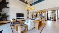 This thoughtfully designed amenity space combines coastal-modern style with a range of functional areas across a single floor. The grand lounge anchors the space with soaring double-height ceilings accented by a deep teal coffered detail, large floor-to-ceiling windows that flood the room with natural light, and dramatic woven rattan pendant lights. Plush white sofas, natural wood furnishings, and a sleek fireplace wall with an integrated TV create a welcoming, resort-like atmosphere. Large-format light tile flooring runs throughout, providing a seamless visual flow between areas. A fully equipped kitchen and bar area features a generous island with quartz countertops, warm wood cabinetry, pendant lighting, and a bold patterned tile backsplash, opening to an adjacent dining space with a dark wood table, woven globe pendant lights, and a striking geometric parquet-style area rug. The game room offers a lively retreat with bold graphic wallcovering, deep blue lounge seating, a billiards table, and a display screen. A dedicated workspace nook, tucked behind a slatted wood partition, provides a quiet area for productivity. Welcoming entryways with black steel-framed glass doors and curated greenery complete the space's polished, resort-inspired character.