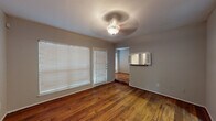 This single-story space is characterized by rich, warm-toned hardwood flooring that flows consistently throughout the main living areas, creating a cohesive and inviting atmosphere. The living area is well-lit by multiple windows fitted with white horizontal blinds, allowing ample natural light to fill the space. A decorative ceiling light fixture with a fan anchors the room, which also features a pass-through opening to an adjacent area, adding an open, connected feel to the layout.

The galley-style kitchen is efficiently designed with white cabinetry lining both sides, dark granite-style countertops, and black appliances including a range and refrigerator. Hardwood flooring continues into the kitchen, maintaining visual continuity throughout.

The bathroom area features a dedicated vanity space with white cabinetry, a dark granite countertop, and a large mirror with overhead lighting, providing a functional and well-appointed grooming area.

The bedrooms offer varied flooring options, with at least one room featuring plush carpeting and a ceiling fan, while another presents hardwood floors and bright windows. Throughout the space, neutral gray wall tones complement the warm flooring, creating a balanced and versatile interior ready for personalization.