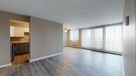Located at 25151 Brookpark Rd in North Olmsted, Ohio, this single-floor residential unit offers a well-organized layout with a blend of updated finishes and functional design. The open living area is bright and spacious, featuring light gray wood-look flooring, neutral taupe walls, and a ceiling fan. A wide wall of vertical blinds covers expansive windows that invite generous natural light and provide outdoor views, complemented by a baseboard heating unit and wall-mounted air conditioner. The kitchen is thoughtfully appointed with two-tone cabinetry — white upper cabinets and slate-blue lower cabinets — paired with light marble-style countertops and a subway tile backsplash. Stainless steel appliances, including a range with over-the-range microwave, complete the space, all set atop matching light wood-look flooring. The bedroom offers plush carpeting, neutral gray walls, a standard closet with paneled doors, and a window with baseboard heat below. The bathroom is neatly finished with tile flooring, a tub-shower combination featuring floor-to-ceiling tile surround, a gray shaker-style vanity with marble-look countertop, a large mirror, and modern light fixtures. The entry area provides a clean transition into the main living space with consistent flooring throughout.