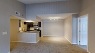 This single-floor space showcases an open-concept layout that seamlessly integrates the kitchen and living area. The kitchen is outfitted with dark espresso cabinetry, stainless steel appliances — including a refrigerator, microwave, and range — and a peninsula countertop with granite-style surfaces that doubles as a breakfast bar. The kitchen features hardwood-style flooring and a tile backsplash, adding texture and contrast to the darker cabinetry.

A half-wall peninsula serves as a natural transition into the carpeted living area, which is finished with neutral-toned carpet and light gray walls. The living area benefits from notably high ceilings, creating an expansive and airy atmosphere. Track lighting mounted near the upper wall provides direct illumination to the living space, complementing the overall brightness of the unit.

The elevated ceiling height in the kitchen zone, visible from the living area, adds architectural interest and contributes to the open, lofty feel of the space. The neutral color palette throughout — soft grays, warm whites, and natural wood tones — creates a cohesive and versatile environment.