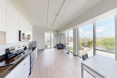 Located on the sixth floor at 169 Canal St in Providence, Rhode Island, Unit 610 is a thoughtfully designed studio that makes efficient use of its intimate footprint. The open-plan living area is the heart of the unit, characterized by clean white walls, light hardwood-style flooring, and floor-to-ceiling windows that flood the space with natural light and frame views of the surrounding Providence cityscape, including nearby greenery and urban architecture. Track lighting runs along the ceiling, providing adjustable illumination throughout. The kitchen is positioned along one wall and features sleek white cabinetry, dark countertops, stainless steel appliances including a refrigerator and dishwasher, and a sink — offering a fully functional culinary space within a compact layout. A partial partition wall subtly defines the sleeping area from the main living and workspace zone. The hallway connects the living space to the full bathroom, which is finished with neutral tones, tile flooring, and includes accessibility-friendly grab bars. The unit's modern, minimalist aesthetic is consistent throughout, making it a practical and contemporary urban living space in the heart of Providence.