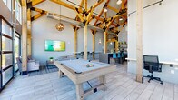Located at 9425 20 Mile Rd in Parker, Colorado, this single-floor amenity space delivers an impressive blend of rustic and modern design. The architectural centerpiece is a dramatic timber-frame ceiling structure, with bold wooden trusses and black metal accents rising to extraordinary heights, filling the space with a sense of grandeur and warmth. Light-toned wood-look flooring runs throughout, complementing the natural materials overhead.

Floor-to-ceiling windows and a full-width glass panel garage door flood the main gathering area with abundant natural light while offering sweeping views of the surrounding Colorado landscape. The garage door opens completely, creating a seamless indoor-outdoor connection to an exterior patio furnished with seating, ideal for year-round enjoyment. A wall-mounted flat-screen TV and comfortable lounge seating anchor one end of the main room, while a full-size billiard table serves as a central recreational focal point, all beneath a statement globe pendant light.

The kitchen area is thoughtfully appointed with dark cabinetry, open shelving, a stainless steel refrigerator, and a large dark-topped island with bar-height seating in modern blue chairs. Decorative wood-panel wall art and patterned wallpaper add texture and personality to the kitchen zone, which also features additional counter-height wall seating for casual dining or work.