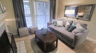 This single-floor space showcases a thoughtfully designed living area that balances comfort and style. The living room is anchored by a plush gray sofa adorned with patterned and teal accent pillows, creating a welcoming focal point. A rich, dark-wood oval coffee table with a lower shelf sits atop a neutral area rug, adding warmth and functionality to the space. Complementing the seating arrangement is a upholstered ottoman, offering additional flexible seating or surface space.

Natural light is a defining feature of this room, streaming in through large floor-to-ceiling windows dressed with vertical blinds and flanked by dark charcoal drape panels, allowing for adjustable light control. The walls are finished in a soft, warm neutral tone that enhances the brightness of the space. Dark hardwood-style flooring grounds the room and contrasts elegantly with the lighter furnishings. A framed wall mirror and a table lamp add finishing touches, contributing to the room's polished, cohesive aesthetic. Overall, the space presents a clean, modern sensibility with a warm and livable character.