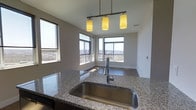 Located at 900 Luxor Lane in Norristown, Pennsylvania, this single-floor residence offers a refined living experience with a thoughtfully designed interior. The kitchen is a standout feature, showcasing elegant granite countertops with a deep stainless steel sink and a sleek pull-down faucet. A modern three-pendant light fixture hangs overhead, casting warm, focused light across the workspace and adding a contemporary aesthetic to the room. The kitchen opens seamlessly toward a bright dining or sitting area, where an impressive arrangement of large windows wraps around the corner, flooding the space with abundant natural light. These expansive windows frame sweeping elevated views of the surrounding Norristown landscape, offering a striking panorama that stretches across the horizon. The neutral color palette throughout — with clean white walls and light flooring — creates a calm, airy ambiance that complements the open layout. The elevated position of the unit enhances the sense of space and openness, making natural light a defining characteristic of the interior. Overall, the residence combines modern finishes with practical design, resulting in a bright and inviting living environment with notable architectural detail and compelling outdoor vistas.
