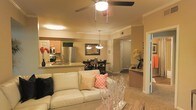 This single-floor residence offers a thoughtfully designed open-concept layout that effortlessly connects the main living areas. The living room anchors the space with a cozy, neutral-toned sofa accented by decorative throw pillows, a ceiling fan with integrated lighting, and crown molding that adds a refined architectural touch. The space flows naturally into a dedicated dining area, highlighted by an elegant pendant light fixture and a dark-toned dining set, perfect for both everyday meals and entertaining. The kitchen is well-appointed with stainless steel appliances, including a full-size refrigerator and range, warm wood cabinetry, and granite-style countertops that extend to a convenient breakfast bar. The open pass-through between the kitchen and living areas enhances the sense of connectivity and spaciousness throughout. Warm ambient lighting and a consistent neutral color palette unify the space, creating a welcoming and comfortable atmosphere. Additional hallway access hints at further private areas beyond the main living zone, rounding out a functional and attractive floor plan designed for comfortable everyday living.