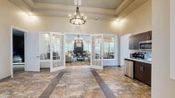 Located at 457 Dutch Meadows Ln in Schenectady, NY, this single-story amenity building offers a thoughtfully designed collection of spaces suited for both relaxation and productivity. The grand entrance lobby immediately impresses with soaring ceilings, an elegant chandelier, and richly toned stone-look tile flooring accented by decorative inlays. French doors open into a welcoming lounge area anchored by a tile-surround fireplace, plush seating, a patterned area rug, and abundant natural light from large windows and glass exterior doors. Adjacent to the lobby is a compact kitchenette outfitted with dark cabinetry, stainless steel appliances, and a sink — ideal for refreshments. A private meeting room features a warm wood round table, upholstered seating, and artwork, while a dedicated office suite provides multiple workstations and a conference table in a bright, professionally appointed setting. The fitness center is divided into two areas: a cardio room with treadmills, ellipticals, and a stationary bike, and a strength area with a full dumbbell rack and floor-length mirror. A private theater room with tiered leather recliner seating and a projection screen rounds out the amenities. Restrooms and dual water fountains ensure full convenience throughout.