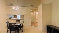 This single-story space is finished in soft, neutral tones throughout, creating a calm and cohesive ambiance. The dining area is a focal point, anchored by a dark wood round table and chairs beneath an elegant chandelier that adds a touch of classic charm. The ceiling height in this area is notably elevated, enhancing the sense of openness. Adjacent to the dining space, a pass-through counter connects to the kitchen, which features a sink, cabinetry, and a tiled backsplash visible through the opening — a practical layout that maintains visual connectivity between the two areas. The flooring transitions from plush, light-colored carpet in the dining and living areas to tile in the hallway and beyond, clearly delineating the functional zones. A wide, white-trimmed hallway extends from the main living area, leading to additional rooms, including what appears to be a bedroom with an en-suite bathroom visible in the distance. The walls are painted in warm beige tones, and recessed lighting in the hallway complements the chandelier, ensuring the space is well-lit throughout.