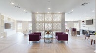 This single-floor community amenity space is thoughtfully designed to encourage gathering and relaxation. The open-plan layout flows seamlessly between distinct functional zones, all unified by light wood-tone flooring installed in a herringbone-influenced pattern and a neutral, warm color palette throughout.

The central lounge area serves as the heart of the space, anchored by a striking floor-to-ceiling decorative lattice screen with an art deco-inspired fan motif. Rich plum-colored armchairs surround a geometric glass and metal coffee table, creating an inviting focal point. Recessed lighting above illuminates the area with a clean, modern glow.

Adjacent to the lounge, a casual dining area features a dark wood table with contemporary grey chairs, positioned near large windows and glass French doors that open to an outdoor patio, flooding the space with natural light and views of the surrounding landscape.

A well-appointed wet bar area includes a white quartz countertop, under-counter refrigeration, a sink, wall-mounted shelving, and a flat-screen TV — ideal for entertaining. High-top bar seating in matching plum tones reinforces the cohesive design language.

A quieter nook with built-in shelving and two neutral-toned accent chairs provides a more intimate retreat within the space. A coffered ceiling detail in the dining zone adds architectural depth, while large windows throughout ensure the space remains bright and connected to the outdoors.