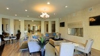 This single-story space showcases a thoughtfully designed open-concept lounge area that exudes a welcoming yet polished ambiance. The main seating area is anchored by a sleek, wall-mounted fireplace framed in textured stone or tile cladding, paired with a flat-screen television mounted above, creating a refined focal point. A statement chandelier with frosted glass shades hangs from the ceiling, complementing recessed lighting throughout to provide a warm, layered glow across the space.

The lounge is furnished with a curated arrangement of neutral-toned sofas and accent chairs accented with blue throw pillows, centered around a woven coffee table. A tall potted succulent adds a natural, organic touch to the contemporary décor. The flooring transitions between hardwood-style planks and area rugs, defining the seating zones while adding textural contrast.

Broad arched openings on either side lead seamlessly into additional areas, including what appears to be a dining or gathering space visible in the background, enhancing the sense of openness and flow. The overall palette of warm beiges, grays, and blues contributes to a calm, modern aesthetic throughout.