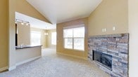 This single-story home showcases an open-concept layout that seamlessly connects the living area to the adjacent kitchen space. The living room is anchored by a striking floor-to-ceiling stacked stone fireplace with a classic black insert, serving as a focal point that adds warmth and texture to the room. Vaulted ceilings rise overhead, enhancing the sense of spaciousness and airiness throughout the main living area.

Large windows allow generous amounts of natural light to fill the space, illuminating the neutral-toned walls and plush wall-to-wall carpeting that runs throughout the living area. The warm beige color palette on the walls complements the natural stone of the fireplace, creating a cohesive and inviting atmosphere.

Adjacent to the living room, the kitchen area is partially visible and features a raised breakfast bar counter with dark cabinetry below, adding a subtle contrast to the lighter surroundings. A decorative chandelier overhead provides additional lighting to this transitional space. The open sightlines between the kitchen and living area make this home well-suited for everyday living and entertaining alike.