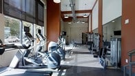 This two-floor fitness facility is designed to support a comprehensive workout experience. The main exercise floor is generously sized, offering ample room for both cardio and strength training. A full lineup of cardio equipment — including elliptical machines, rowing machines, and treadmills — is arranged along the perimeter, benefiting from large windows that flood the space with natural light. On the opposite side, a dedicated strength-training zone features cable machines and resistance equipment to accommodate a variety of exercise routines.

Architecturally, the space is defined by soaring ceilings with exposed industrial-style ductwork and bold terracotta-red accent columns that create a dynamic, energetic atmosphere. Pendant lighting and a ceiling fan are suspended overhead, ensuring the space remains well-lit and well-ventilated throughout. The flooring transitions between hard surface and rubberized mat sections, thoughtfully delineating different functional workout zones.

Large roller shades on the windows allow for flexible light control, making the space adaptable for different times of day. The overall design balances an industrial aesthetic with functional practicality, resulting in an inviting and motivating environment for fitness enthusiasts.