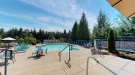 This outdoor amenity space centers around a generously sized swimming pool with clear, turquoise water that reflects the open sky above. The pool is framed by a wide, light-toned concrete deck that provides ample room for movement and relaxation. Surrounding the pool are numerous lounge chairs and chaise lounges in teal and neutral tones, neatly arranged to offer poolside comfort and sun exposure throughout the day. Small seating clusters with chairs and tables are also positioned on the deck, providing casual gathering spots. Metal handrails lead down steps from an elevated section of the deck, indicating a slight change in elevation that adds dimension to the layout. The perimeter of the space is enclosed by a dark metal fence, offering both safety and a clean visual boundary. Tall evergreen trees and lush greenery form a natural backdrop that surrounds the area, providing privacy and a sense of seclusion. The overall atmosphere is open, airy, and well-maintained, with abundant natural light and a welcoming outdoor setting suited for leisure and recreation.