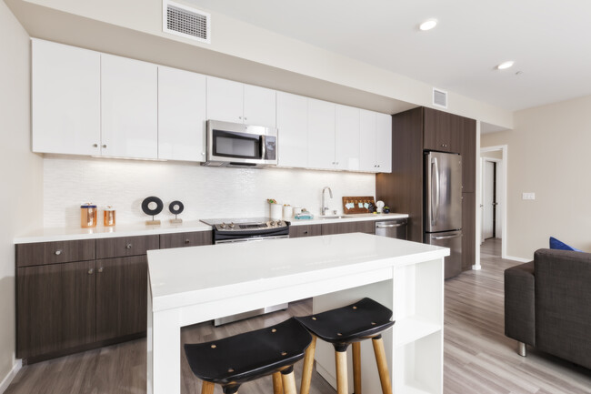 Open concept kitchen with stainless steel appliances, white and brown cabinetry, quartz stone countertop, ceramic tile backsplash, island table and hard surface flooring - Avalon Dogpatch