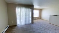 This single-floor space presents a straightforward, functional layout centered around a generously sized living area. The room is finished with wall-to-wall carpet in a neutral gray tone, providing a soft underfoot feel that extends throughout the open floor plan. Neutral-toned walls complement the flooring and contribute to a clean, understated aesthetic.

A prominent sliding glass door dominates one wall, dressed with full-length vertical blinds that offer both privacy and light control. The door provides access to an outdoor area, allowing natural light to pour into the interior. Adjacent to the sliding door, a secondary window further brightens the space, enhancing the airy feel of the room.

A baseboard heating unit is positioned beneath the secondary window, indicating a practical heating solution integrated into the room's perimeter. The flat ceiling maintains a consistent height throughout, reinforcing the space's clean lines and simple design. The overall ambiance is neutral and move-in ready, offering a blank canvas well-suited for a variety of interior arrangements and personal styles.