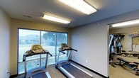 This single-floor fitness room is designed with functionality and practicality in mind, offering a dedicated space for cardiovascular exercise. The room features multiple treadmills positioned to take full advantage of the large windows along one wall, which flood the space with natural light and provide views of the surrounding outdoor greenery — an inviting backdrop that enhances the workout experience. The flooring is a durable, low-maintenance surface well-suited for a high-traffic exercise environment. Overhead, bright flush-mounted fluorescent lighting panels ensure the room remains well-lit at all hours. The walls are finished in neutral tones — a warm beige accent wall paired with clean grey surfaces — creating a calm, focused atmosphere. The layout is straightforward and efficient, maximizing usable floor space for equipment. Horizontal blinds on the windows offer adjustable light and privacy control as needed. The overall ambiance is clean, modern, and utilitarian, making it a practical and welcoming space for anyone looking to maintain an active lifestyle.