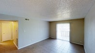This single-floor space offers a clean, functional layout with a welcoming open feel. The main living area is finished with neutral-toned tile flooring that provides durability and easy maintenance, while the walls are painted in a soft, warm beige that contributes to a bright and airy atmosphere. A textured popcorn ceiling adds a classic architectural detail overhead, and a central HVAC vent ensures comfortable climate control throughout the space. A prominent sliding glass door, dressed with vertical blinds, serves as the focal point of the room, allowing abundant natural light to flood the interior. The door opens to an outdoor balcony or patio area, offering a seamless connection between the interior living space and the outdoors. White baseboards provide a crisp contrast against the tile flooring, lending a tidy, polished finish to the room. The space is move-in ready, with multiple electrical outlets and switch plates thoughtfully positioned along the walls for convenience. Overall, the layout is straightforward and versatile, well-suited for a variety of furniture arrangements and uses.
