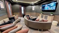 This single-floor lounge or media room is thoughtfully designed for communal relaxation and entertainment. The centerpiece of the space is a generously sized curved sectional sofa upholstered in a warm taupe fabric, adorned with an array of mauve, blush, and charcoal accent pillows that add texture and visual interest. Additional curved bench seating lines the perimeter of the room, maintaining the space's cohesive, circular layout and maximizing seating capacity for group gatherings.

A large wall-mounted flat-screen television serves as the focal point for viewing, positioned prominently on one wall alongside a low credenza with display shelving. The room's walls are finished in a deep charcoal tone, complemented by a two-tone wainscoting detail that adds architectural character. Recessed lighting within a tray ceiling provides ambient illumination, while a curtained window allows for natural light and privacy control. Framed artwork adds a decorative touch to the walls. The patterned carpeted flooring ties together the room's warm, neutral color palette, creating an inviting and sophisticated atmosphere throughout.