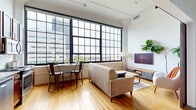This loft-style residence blends industrial character with modern comfort across a single thoughtfully designed floor. The entrance leads down a clean corridor with warm hardwood flooring that carries throughout the main living areas. The combined kitchen and living room serves as the heart of the home, anchored by a dramatic wall of steel-frame, grid-pane windows that flood the space with natural light and frame sweeping city skyline views. The kitchen features warm wood cabinetry, stainless steel appliances, and ample counter space, while the living area offers generous room for lounging and dining. Exposed ceiling elements and industrial pendant lighting reinforce the loft aesthetic. The bedroom is a cozy, well-defined retreat accessed via a stylish sliding barn door, offering direct access to a dedicated closet. The full bathroom is finished with tile flooring, a granite-topped wood vanity, a large framed mirror, and a glass-enclosed shower. Additional conveniences include in-unit laundry. The bright, airy ambiance, city views, and cohesive industrial-modern design make this a distinctive urban living space.