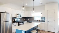 This contemporary multi-floor residence is thoughtfully designed with modern finishes and a cohesive aesthetic throughout its three levels. The kitchen serves as a standout centerpiece, featuring crisp white cabinetry with matte black hardware, a bold teal island topped with a white quartz countertop, and a textured dark tile backsplash. Stainless steel appliances, a deep undermount sink, and a pair of oversized globe pendant lights complete this highly functional and visually striking space. Light wood-tone flooring flows seamlessly through the main living areas, enhancing the bright, airy atmosphere created by abundant natural light from multiple windows. The living room offers a relaxed, open feel with a sliding glass door that connects to an outdoor balcony, inviting views of the surrounding landscape. Bedrooms are carpeted for comfort, with large windows that frame outdoor views and allow generous natural light. A well-appointed bathroom features classic white subway tile surrounding a soaking tub with an oil-rubbed bronze fixture, adding a refined contrast to the otherwise clean, neutral palette. The entryway provides a welcoming transition into the home with clean lines and a slatted bench. Throughout, the design balances modern style with practical livability across all three floors.