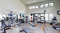 This fitness center is designed to support a comprehensive workout experience in a bright, open environment. The space immediately impresses with its dramatically high ceilings and an array of clerestory windows that flood the interior with natural light, creating an energizing and airy atmosphere. The flooring features a modern hardwood-style surface accented with bold orange and gray stripe detailing, adding a dynamic visual element to the space.

The strength training area is well-appointed, featuring a full dumbbell rack with a wide range of weights, an adjustable workout bench, and large wall-mounted mirrors that enhance the sense of space while allowing users to monitor their form. A multi-functional cable and pulley machine provides versatile resistance training options for users at all fitness levels.

A dedicated functional training zone houses a medicine ball rack stocked with various weighted balls, along with kettlebells and other accessories for dynamic and circuit-style workouts. An interactive fitness console provides guided training options, adding a modern, tech-forward dimension to the facility. The clean white walls, organized equipment layout, and generous floor space all contribute to a professional, gym-quality environment that is both practical and inviting.