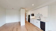 Located at 1341 N Killingsworth St in Portland, Oregon, this thoughtfully designed studio unit offers an efficient and modern single-floor layout. The open living area is anchored by light hardwood flooring throughout, crisp white walls, and recessed lighting that keeps the space bright and airy. A sliding glass door and flanking windows flood the room with natural light and provide access to a private balcony overlooking lush greenery.

The kitchen is well-appointed with white flat-front cabinetry extending to the ceiling for maximum storage, a clean white countertop, an undermount sink with a matte black faucet, a black refrigerator, built-in microwave, electric cooktop, and a dishwasher — all finished with coordinating black hardware. A stacked washer and dryer unit is neatly integrated behind a partition wall, offering the convenience of in-unit laundry without sacrificing living space.

A hallway connects to the full bathroom, which continues the clean, modern aesthetic with white cabinetry, a floating vanity with black hardware, a full soaking tub and shower combination with matte black fixtures, and matching hardwood-style flooring. The consistent design language throughout — neutral tones, black accents, and light wood floors — gives the entire unit a cohesive and contemporary feel.