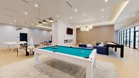 Located at 632 Newark Avenue in Jersey City, NJ, this expansive single-floor amenity lounge is a thoughtfully designed recreational and social space. The open-plan layout spans a generous footprint, offering a seamless flow between distinct activity zones. Light hardwood flooring runs throughout, complemented by a neutral palette of whites and warm wood tones that create a clean, contemporary ambiance.

The game area is a central highlight, featuring a pool table with a teal felt surface, a full-length shuffleboard table, a foosball table, and a ping pong table — catering to a wide range of recreational activities. A dedicated lounge zone offers plush blue sectional sofas, accent chairs with woven detailing, and area rugs that define the seating area, all anchored by a textured stone fireplace with a mounted flat-screen TV above.

Vertical wood slat wall panels add warmth and architectural texture, while globe and industrial pendant lighting fixtures provide layered illumination. Large black-framed floor-to-ceiling windows and glass doors flood the interior with natural light and offer views into adjacent spaces. A tray ceiling with recessed lighting adds dimension to the layout. The overall design balances a modern aesthetic with a welcoming, community-oriented atmosphere.