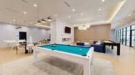 This 2,240 square foot single-floor space located at 634 Newark Ave, Jersey City, NJ, is a meticulously designed communal area that seamlessly blends functionality with style. The open-plan layout is characterized by its modern aesthetic, featuring light wood flooring and a sophisticated neutral color palette. 

The space is divided into distinct zones, each serving a specific purpose. A prominent game area includes a pool table and shuffleboard table, providing entertainment options for residents. Comfortable lounge spaces are scattered throughout, featuring plush sofas and armchairs in muted tones, perfect for relaxation or casual conversations.

The room's ambiance is enhanced by its thoughtful lighting design. Recessed ceiling lights are complemented by pendant lamps and indirect lighting, creating a warm and inviting atmosphere. Large windows allow ample natural light and offer views of the surrounding area.

A notable feature is the adjoining conference room, visible through glass partitions, which adds versatility to the space. Modern amenities include wall-mounted TVs and contemporary artwork, contributing to the overall upscale feel.

The attention to detail is evident in the textured wall treatments, including wood paneling and decorative wall coverings. This multifunctional space is designed to foster community interaction while providing areas for both active recreation and quiet relaxation.