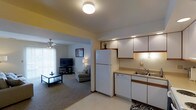 This single-floor unit offers an open and functional layout that seamlessly connects the kitchen and living area. The kitchen is outfitted with white cabinetry featuring wood-trimmed accents, a full-size white refrigerator, a dishwasher, and a double-basin sink positioned beneath under-cabinet lighting. The countertops provide ample prep space, and the cabinetry extends to the ceiling, maximizing storage. Recessed and accent lighting keeps the kitchen well-illuminated.

The adjoining living area is defined by neutral carpet flooring and is accessible through an open archway. A ceiling fan with a light fixture provides both airflow and ambient lighting, while vertical blinds along a large window allow for adjustable natural light. The living space comfortably accommodates a media console with a flat-screen television and seating furniture, lending a relaxed residential atmosphere. Warm yellow accent walls add a touch of color to the otherwise neutral palette. The overall design is practical and cohesive, making efficient use of the available space throughout the open floor plan.