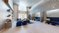 Located at 3240 W Harmon Ave in Las Vegas, Nevada, this elegantly designed lobby lounge offers a refined and welcoming atmosphere across a generously proportioned single-floor layout. The space is defined by dramatically soaring double-height ceilings that amplify the sense of openness, complemented by large floor-to-ceiling windows dressed in sheer white curtains that flood the interior with natural light and frame views of the Las Vegas skyline, including glimpses of the iconic Strip.

The main lounge area features a curvaceous sectional sofa in soft grey, accented by bold navy blue armchairs and intimate booth-style seating separated by sleek slatted wood and metal partitions, creating semi-private conversation zones. Large-format light tile flooring runs throughout, grounding the contemporary aesthetic.

Toward the rear, a distinct social hub showcases a dramatic textured feature wall in rich charcoal tones, illuminated by a row of gold pendant lights that cast a warm glow over a bar counter, communal dining table, and additional lounge seating. Curated wall art and lush greenery add personality and warmth to the space.

The overall design palette — neutral tones of white, grey, and beige layered with deep navy blue and gold accents — projects a modern, upscale character well-suited to its vibrant Las Vegas location.