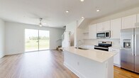 This well-designed two-story home offers a clean, contemporary aesthetic with a thoughtful layout across two floors. The ground floor opens with an entrance that leads into a bright, open-concept kitchen and living area. The kitchen is equipped with stainless steel appliances—including a range, microwave, and refrigerator—white shaker-style cabinetry, a white quartz waterfall island with an integrated sink, and a subway tile backsplash. The living area features light hardwood-style flooring, recessed lighting, a ceiling fan, and large sliding glass doors that invite natural light and provide outdoor views. An attached garage with an automatic opener completes the ground floor. The staircase connects to the upper level, where three bedrooms are located, each featuring the same warm hardwood-style flooring, ceiling fans, and windows that bring in natural light. The primary bedroom includes an en-suite bathroom and closet access. The bathrooms are finished with neutral tile, including a walk-in glass-enclosed shower with large-format tile and a built-in niche. Two additional closets provide ample storage. The home's bright white walls, consistent flooring, and modern fixtures create a cohesive and inviting atmosphere throughout.