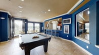 This single-floor community amenity space is designed for social gathering and recreation, characterized by bold cobalt blue accent walls, crown molding, and large-format tile flooring throughout. The main game and lounge area serves as the centerpiece, anchored by a full-size pool table and complemented by a seating arrangement of brown leather sofas and armchairs with decorative pillows. Recessed lighting and cinema-themed wall art add personality and visual interest to the space. Large windows dressed with horizontal blinds and floor-length curtains line the walls, flooding the interior with abundant natural light and offering views of the surrounding greenery. A dedicated dining nook features a round table with modern chairs, positioned beside multiple windows to maximize the connection to the outdoors. The fully equipped kitchen is outfitted with warm wood cabinetry, dark granite countertops, stainless steel appliances including a refrigerator and microwave, and a tiled backsplash. A peninsula with a bold blue fascia accommodates bar-height seating with sleek metal stools, making it ideal for casual gatherings. Pendant cage lights hang above the kitchen area, adding an industrial-chic touch to the overall warm and energetic ambiance of the space.