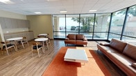 This single-floor commercial space is designed as a contemporary lounge and break room, combining comfort and functionality in an open, well-lit layout. The most striking architectural feature is the expansive floor-to-ceiling glass curtain wall that wraps around a corner of the space, flooding the interior with natural light and offering unobstructed views of a waterfront setting framed by mature trees. Wood-look tile flooring runs throughout, complementing the neutral, warm-toned walls and drop-panel ceiling with recessed lighting.

The lounge area is anchored by rich brown leather sofas arranged around a white coffee table on an orange area rug, creating an inviting, relaxed seating zone. A wall-mounted flat-screen television adds a functional media element to this area. Transitioning further into the space, a casual dining section features multiple round white tables paired with mid-century modern wood-and-black chairs, offering flexible seating arrangements. This area is served by built-in cabinetry with a countertop along one wall, providing practical storage and workspace. Vending machines offering snacks and beverages are conveniently positioned nearby, rounding out the amenities. Together, these elements create a bright, comfortable, and fully equipped communal space suited for relaxation and informal gatherings.