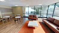 This single-floor commercial space is designed as a contemporary lounge and break room, combining comfort and functionality in an open, well-lit layout. The most striking architectural feature is the expansive floor-to-ceiling glass curtain wall that wraps around a corner of the space, flooding the interior with natural light and offering unobstructed views of a waterfront setting framed by mature trees. Wood-look tile flooring runs throughout, complementing the neutral, warm-toned walls and drop-panel ceiling with recessed lighting.

The lounge area is anchored by rich brown leather sofas arranged around a white coffee table on an orange area rug, creating an inviting, relaxed seating zone. A wall-mounted flat-screen television adds a functional media element to this area. Transitioning further into the space, a casual dining section features multiple round white tables paired with mid-century modern wood-and-black chairs, offering flexible seating arrangements. This area is served by built-in cabinetry with a countertop along one wall, providing practical storage and workspace. Vending machines offering snacks and beverages are conveniently positioned nearby, rounding out the amenities. Together, these elements create a bright, comfortable, and fully equipped communal space suited for relaxation and informal gatherings.