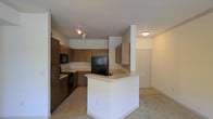 This single-story residence in Roanoke, TX presents a practical and functional layout across its one level. The floor plan, as revealed by the 3D model, features multiple interconnected rooms arranged in an L-shaped configuration, offering a comfortable sense of space throughout.

The kitchen is a notable focal point, equipped with dark wood cabinetry that provides ample storage, light-toned countertops, and a suite of black appliances including a refrigerator, range, and built-in microwave. A peninsula extends from the kitchen, creating a natural boundary between the cooking area and the adjacent living space while also offering additional counter space. Track lighting above the kitchen ensures the area is well-lit.

The living area is generously proportioned, with neutral-toned carpet flooring that extends through much of the home, contributing to a cohesive and understated aesthetic. White walls throughout the space keep the interior feeling bright and open. Additional rooms branch off from the central living space, each accessible through standard doorways. The overall layout is straightforward, making it easily adaptable for a variety of uses.