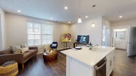This well-designed single-floor unit offers a cohesive, modern aesthetic with clean lines and a bright, neutral color palette throughout. The open-concept layout seamlessly connects the kitchen, dining, and living areas, creating a spacious and functional flow. Rich dark hardwood flooring anchors the main living spaces, providing a striking contrast against the crisp white walls and trim.

The kitchen is a standout feature, showcasing white shaker-style cabinetry, a large central island with a white quartz countertop and seating for two, stainless steel appliances, and elegant pendant lighting overhead. The entry area features a transom window above the front door, allowing additional natural light to filter in.

The open living area is bright and inviting, with large windows dressed in white plantation blinds that offer ample natural light. The space is well-suited for both relaxing and entertaining.

The bedroom is a comfortable retreat, finished with plush carpeting, a ceiling fan, and multiple windows that bring in generous natural light and outdoor views.

The bathroom is thoughtfully finished with a white shaker-style vanity, white quartz countertop, well-lit mirror, and dark hardwood-style flooring that echoes the main living areas. Throughout the unit, recessed lighting and carefully chosen fixtures contribute to a polished, contemporary atmosphere.