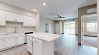 This well-appointed single-floor unit offers a thoughtfully designed open-concept layout that seamlessly connects the kitchen, dining, and living areas. The kitchen is a standout feature, showcasing crisp white shaker cabinetry, a large center island with a bright white countertop, stainless steel appliances including a refrigerator, range, microwave, and dishwasher, and durable light-toned hardwood-style flooring that extends throughout the main living areas. Recessed lighting and a ceiling fan with a light fixture enhance the living space, which opens to a private balcony through sliding glass doors, offering pleasant outdoor views of surrounding greenery. The bedrooms are carpeted for comfort and feature generous closet space with built-in wire shelving and sliding doors, along with windows that bring in ample natural light. The bathroom is clean and functional, with a white vanity, a tub-shower combination, and a window providing natural light. A dedicated in-unit laundry room includes full-size washer and dryer units alongside wire shelving for additional storage. The overall aesthetic is modern and neutral, with warm beige tones, consistent trim details, and an airy, move-in-ready ambiance throughout.