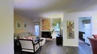 This single-floor space offers a thoughtfully designed interior that balances comfort with artistic flair. The living area serves as the centerpiece of the home, showcasing rich, dark hardwood flooring that anchors the room with warmth and sophistication. A classic brick fireplace provides a focal point, flanked by windows dressed with woven roman shades that allow natural light to filter in while maintaining privacy. The generous window placement ensures the space feels bright and connected to the greenery visible outside. Vibrant, large-scale artwork adorns the walls, adding bold splashes of color that complement the neutral white walls and ceiling. The living area is furnished with a curated mix of seating, including a floral-patterned accent chair and a clean-lined sofa, creating an inviting and eclectic aesthetic. A tall arching floor lamp and a sleek dark media console round out the space, blending functional elements with decorative ones. The open layout promotes a natural flow throughout the area, making the space feel expansive and airy. Overall, the interior strikes a balance between artistic expression and everyday livability, offering a welcoming environment with distinctive character.