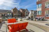 Located in Atlanta, GA (30324), this thoughtfully designed outdoor courtyard serves as a central amenity space within a multi-story residential community. The grounds are anchored by a mix of concrete and decorative paver pathways that curve gracefully through the space, creating a welcoming flow between distinct activity areas.

A dedicated grilling station features a built-in stainless steel gas grill set atop a stone-faced surround, accompanied by an outdoor dining table and seating — ideal for casual gatherings. Nearby, a cozy outdoor fireplace flanked by Adirondack chairs provides a relaxed focal point for cooler evenings, with views of the surrounding neighborhood adding to the open-air ambiance.

Tiered stone retaining walls define landscaped garden beds filled with ornamental grasses, shrubs, and seasonal plantings, lending a natural, layered aesthetic to the hardscape. A lounge area with chaise seating offers a spot for relaxation beneath open skies.

Accessibility is a clear priority, with ADA-compliant ramps and metal handrails integrated alongside stair access points. The surrounding multi-story building facades — clad in a combination of brick and neutral siding — frame the courtyard and contribute to its sheltered, community-oriented character. The overall design balances functionality and comfort in a well-maintained outdoor environment.