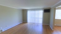 This single-floor space offers a clean and functional layout with a welcoming, understated aesthetic. The main living area is highlighted by warm hardwood flooring that runs throughout, reflecting natural light and adding a sense of warmth to the interior. A large sliding glass door dressed with vertical blinds serves as a prominent architectural feature, flooding the room with abundant natural light and providing direct access to the outdoors. Crown molding along the ceiling adds a subtle decorative touch, lending the space a polished, finished look. The walls are painted in a soft, neutral tone that complements the light wood flooring and contributes to an open, airy feel. A baseboard heating system runs along the perimeter of the room, ensuring efficient and consistent warmth throughout the colder months. A wall-mounted air conditioning unit is also present, offering climate control year-round. The overall ambiance is bright, practical, and move-in ready, making it a versatile space well-suited for a variety of uses and furnishing styles.