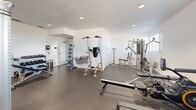 Located at 3480 Todos Santos St in Santa Fe, New Mexico, this fitness center offers a well-equipped exercise space designed to support a comprehensive workout routine. The room features clean white walls, recessed lighting, and durable rubber flooring that provides both safety and comfort during exercise.

The cardio section is outfitted with multiple treadmills, elliptical machines, and a stationary spin bike, all positioned near windows fitted with horizontal blinds that allow natural light to fill the space. A multi-function cable machine by Hoist anchors the strength training area, complemented by a power tower for bodyweight exercises. Additional strength equipment includes adjustable weight benches and a full rack of dumbbells organized on a tiered storage rack, offering a wide range of resistance options.

Stability and accessory training needs are addressed with a collection of medicine balls and stability balls neatly stored along one wall. A water fountain is conveniently mounted near the entry area, alongside a hand sanitizer dispenser and waste receptacle. The space is served by a ceiling ventilation system and recessed can lights that ensure consistent air circulation and visibility throughout. The layout is thoughtfully organized, allowing users to move freely between equipment zones, making this fitness center a functional and efficient facility for residents at this Santa Fe location.