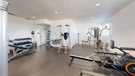 Located at 3480 Todos Santos St in Santa Fe, New Mexico, this fitness center offers a well-equipped exercise space designed to support a comprehensive workout routine. The room features clean white walls, recessed lighting, and durable rubber flooring that provides both safety and comfort during exercise.

The cardio section is outfitted with multiple treadmills, elliptical machines, and a stationary spin bike, all positioned near windows fitted with horizontal blinds that allow natural light to fill the space. A multi-function cable machine by Hoist anchors the strength training area, complemented by a power tower for bodyweight exercises. Additional strength equipment includes adjustable weight benches and a full rack of dumbbells organized on a tiered storage rack, offering a wide range of resistance options.

Stability and accessory training needs are addressed with a collection of medicine balls and stability balls neatly stored along one wall. A water fountain is conveniently mounted near the entry area, alongside a hand sanitizer dispenser and waste receptacle. The space is served by a ceiling ventilation system and recessed can lights that ensure consistent air circulation and visibility throughout. The layout is thoughtfully organized, allowing users to move freely between equipment zones, making this fitness center a functional and efficient facility for residents at this Santa Fe location.