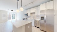This thoughtfully designed unit in Elmhurst, Illinois presents a contemporary aesthetic defined by clean lines, neutral tones, and light-toned hardwood flooring throughout the main living areas. The open-concept kitchen serves as a central feature, showcasing white shaker cabinetry extending to the ceiling, a subway tile backsplash, quartz countertops, and a full suite of stainless steel appliances including a gas range, built-in microwave, dishwasher, and side-by-side refrigerator. A kitchen island with pendant globe lighting adds both functionality and visual interest, while track lighting overhead provides practical illumination.

The living area is bright and airy, benefiting from large sliding glass doors that open onto a private balcony with urban views of the surrounding Elmhurst streetscape. Natural light flows generously through the space. The bedroom features plush carpeting, a tall narrow window framing outdoor views, and direct access to a spacious walk-in closet with multi-tier shelving and hanging rods. The bathroom is finished with a white vanity, tub-shower combination with tiled surround, and black accent hardware, maintaining the unit's cohesive modern palette.