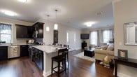 This single-floor space showcases an open-concept layout that integrates the kitchen and living area into one cohesive, flowing environment. The kitchen is outfitted with rich, dark espresso cabinetry that extends to the upper walls, providing ample storage while creating a bold visual contrast against the light-toned walls. The countertops feature striking granite with natural veining, extending into a peninsula breakfast bar complete with dark wood bar stools — an ideal spot for casual dining or entertaining.

Pendant lighting in frosted white glass hangs above the peninsula, complementing the flush-mount ceiling fixtures throughout the space. Crown molding adds a refined architectural detail along the ceiling line, lending a polished finish to the open layout.

The kitchen flows naturally into the living area, where rich hardwood flooring runs continuously underfoot, anchored by a neutral area rug. The living space is bright and welcoming, drawing natural light through two windows dressed with sheer curtains. A wall-mounted flat-screen TV, a cushioned sofa, and a dark wood coffee table complete the comfortable yet functional arrangement. A white paneled entry door with glass accents adds charm to the space.