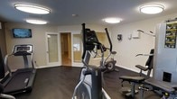 This fitness center offers a dedicated space for exercise and wellness, thoughtfully designed to accommodate a range of workout needs. The room features dark hardwood-style flooring that contrasts cleanly with the light-colored walls, creating a functional and visually grounded environment. Flush-mount ceiling light fixtures provide bright, even illumination throughout the space, ensuring a well-lit area for safe exercise at any time of day.

The facility is equipped with key cardio machines, including an elliptical trainer and a stationary bike, positioned to allow comfortable use without crowding. A weight bench is also present, supporting strength training alongside the cardio options. Wall-mounted accessories, such as a sanitizing station and informational panels, reflect attention to hygiene and user convenience.

A frosted-glass door leads to an adjacent area, adding a sense of openness and connectivity to the broader facility. The overall layout is practical and uncluttered, maximizing usable floor space while maintaining clear pathways between equipment. The neutral color palette and clean finishes contribute to a calm, focused atmosphere well-suited to physical activity. This fitness center serves as a comprehensive amenity space designed to meet everyday exercise needs efficiently.