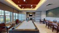 This thoughtfully designed single-floor amenity space combines modern aesthetics with versatile functionality across several distinct zones. The game room serves as a centerpiece, featuring a full-size billiards table beneath a striking tray ceiling finished in a bold terracotta red, accented by cove lighting and a geometric pendant fixture. A lounge area beyond is framed by frosted glass sliding barn doors and decorated with a bold chevron-patterned accent wall, complemented by contemporary curved seating arranged around a flat-screen television.

The kitchen is outfitted with stainless steel appliances, including a full-size refrigerator and built-in wall oven, set against a rich red tile backsplash. Dark wood cabinetry with glass-front uppers lines the walls, while a large island with a white countertop and leather-upholstered bar seating sits beneath a pair of oversized circular pendant lights, creating an inviting space for gathering.

A dedicated café-style dining nook features a continuous built-in bench with red upholstered cushions set against a sculptural three-dimensional wave-textured teal wall panel, paired with wooden tables and patterned side chairs. A secondary lounge area with exposed wood ceiling beams, textured wallcovering, and patterned area rugs provides a quieter retreat. Light tile flooring flows consistently throughout, unifying the space's contemporary design.