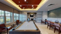This thoughtfully designed single-floor amenity space combines modern aesthetics with versatile functionality across several distinct zones. The game room serves as a centerpiece, featuring a full-size billiards table beneath a striking tray ceiling finished in a bold terracotta red, accented by cove lighting and a geometric pendant fixture. A lounge area beyond is framed by frosted glass sliding barn doors and decorated with a bold chevron-patterned accent wall, complemented by contemporary curved seating arranged around a flat-screen television.

The kitchen is outfitted with stainless steel appliances, including a full-size refrigerator and built-in wall oven, set against a rich red tile backsplash. Dark wood cabinetry with glass-front uppers lines the walls, while a large island with a white countertop and leather-upholstered bar seating sits beneath a pair of oversized circular pendant lights, creating an inviting space for gathering.

A dedicated café-style dining nook features a continuous built-in bench with red upholstered cushions set against a sculptural three-dimensional wave-textured teal wall panel, paired with wooden tables and patterned side chairs. A secondary lounge area with exposed wood ceiling beams, textured wallcovering, and patterned area rugs provides a quieter retreat. Light tile flooring flows consistently throughout, unifying the space's contemporary design.