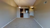 This single-floor space is defined by its vaulted ceilings, which lend an airy, expansive feel to the open-concept layout. The living area features neutral-toned carpet flooring and warm gray walls, creating a clean and welcoming atmosphere. A pendant light hangs prominently from the peak of the vaulted ceiling, serving as a central design element, while a track lighting fixture provides additional illumination toward the kitchen area.

The kitchen is equipped with dark espresso-toned cabinetry, stone-look countertops, and stainless steel appliances, including a refrigerator. A peninsula counter separates the kitchen from the living space, offering a casual seating or serving area and maintaining visual connectivity between the two zones. The kitchen itself features hard flooring, contrasting with the carpeted living area.

A recessed entryway with a white door is visible to the right, providing a defined point of entry. The overall color palette — warm grays, dark wood tones, and white trim — lends the space a cohesive and modern aesthetic. The layout is efficient and well-organized, with distinct functional zones flowing naturally into one another.